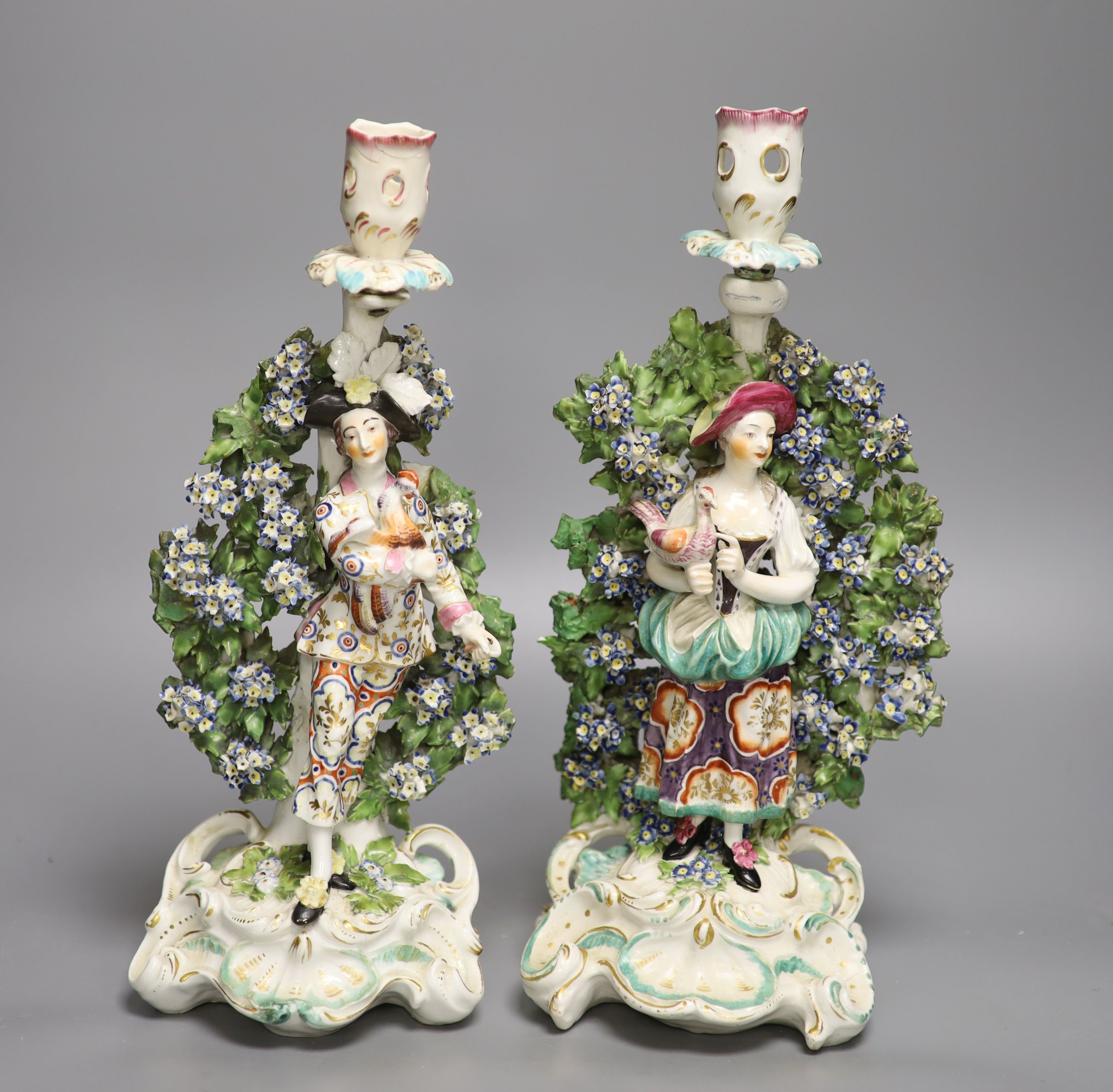 A pair of Derby candlestick figures of the Italian Farmer and wife, he holding a rooster, and her a chicken standing on a rococo scroll